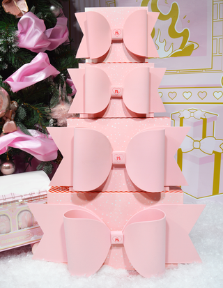 Stack of pink P.Louise gift wrap boxes with bows displayed against a Christmas-themed backdrop with decorations.