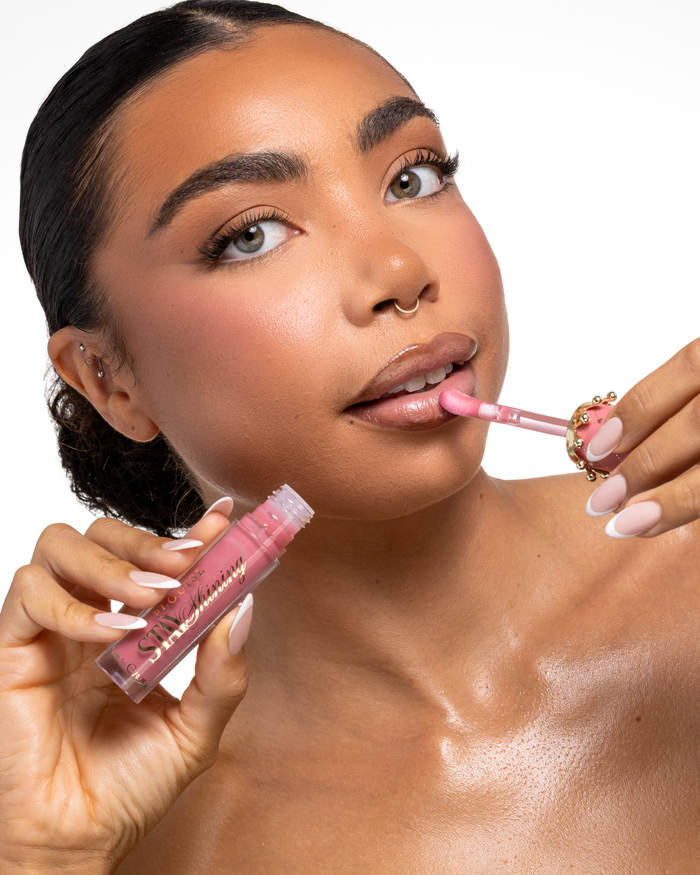 Lähikuva naisesta, joka levittää vaaleanpunaista P.Louise Stay Shining Lip Oil nestemäistä huulituotetta naarasjalka-applikaattorilla pitäen putkea toisessa kädessään puhtaan valkoista taustaa vasten.