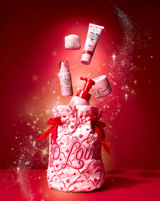 Vaaleanpunainen jouluinen P.Louise-laukku, jossa on punaiset juhlarusetit ja juhlava kuviointi sekä vartalonhoitotuotteet punaisella pohjalla
