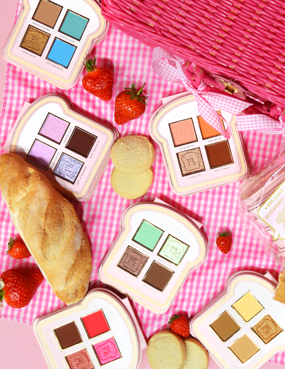 Sarja mini-sandwich-tyylisiä luomiväripaletteja, joissa on erilaisia ​​meikkivärejä vaaleanpunaisella gingham-pinnalla, mukana mansikoita ja pala leipää.