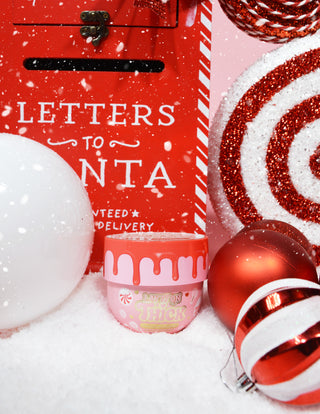 Purkki P.Louise Lay It On Thick Body Butter Frosted Peppermint -versiossa, jonka ympärillä on juhla-aiheisia koristeita, kuten jouluvaloja ja lahjoja.