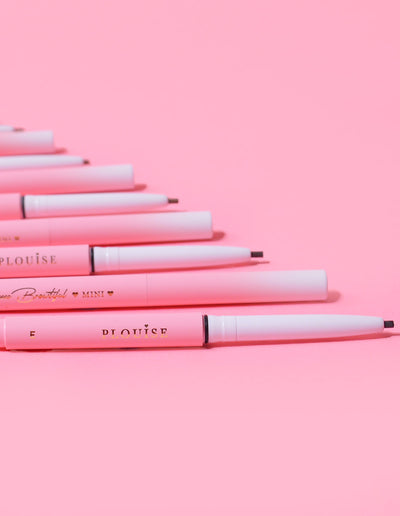 Kokoelma P.Louise Mini Eyebrow Pencils -kyniä, jotka on asetettu riviin ja tuotemerkki näkyy kynien rungossa vaaleanpunaista taustaa vasten